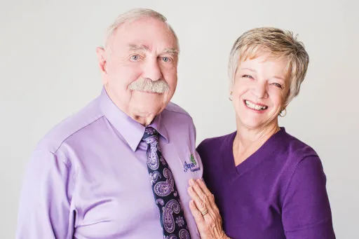 Man in a lilac button-down shirt with tie posing next to a woman, his wife, in a royal purple top - both against a light gray background.